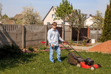 Young man in sunglasses, jeans and shirt mows the grass on the lawn in the village in the summer.