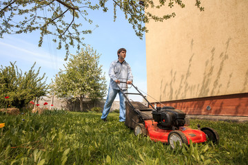 Young man in sunglasses, jeans and shirt mows the grass on the lawn in the village in the summer.