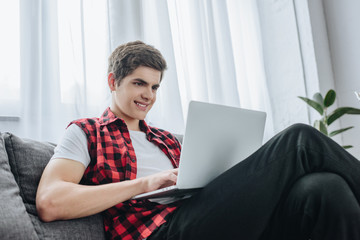 smiling teen boy typing on laptop while sitting on sofa
