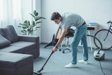 male teenager cleaning floor near sofa with vacuum cleaner