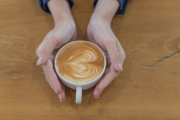 Female hands holding a cup of coffee with foam over wooden table, top view