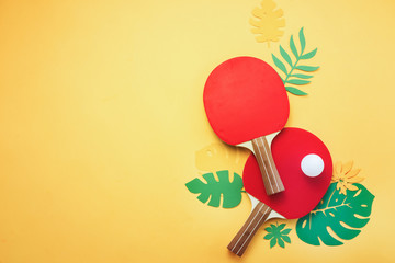 Ping pong rackets and ball with tropical leaves. Summer sports and recreation flat lay on a sunny yellow background with copy space.