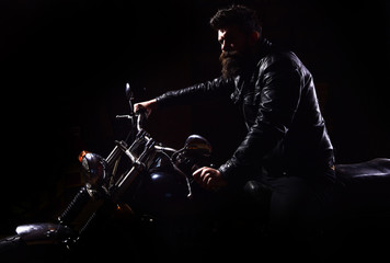 Man with beard, biker in leather jacket sitting on motor bike in darkness, black background. Night racer concept. Macho, brutal biker in leather jacket riding motorcycle at night time, copy space.