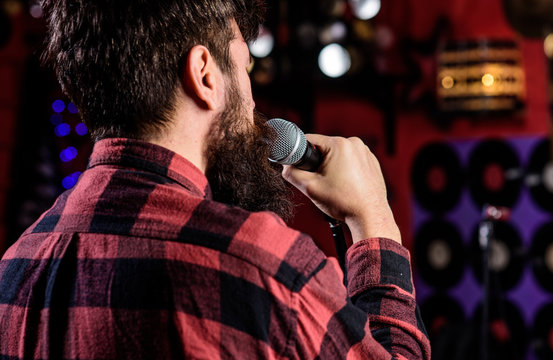 Musician with beard singing song in karaoke, rear view. Rock singer concept. Guy likes to sing in dark karaoke hall. Man in checkered shirt holds microphone, singing song, karaoke club background.