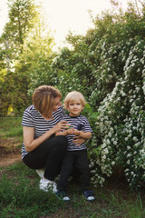 Mother and son in striped T-shirts. Woman hugs and talks to boy. They stand in park by flowering bush in spring. Family time