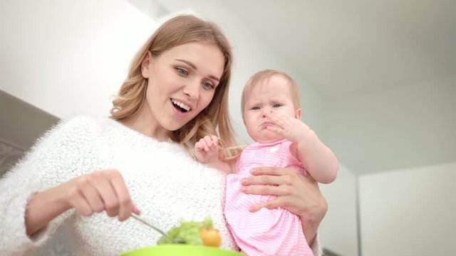 Young mother cooking with baby on hands. Close up of woman holding child on kitchen. Happy mom preparing fresh meal for baby dinner. Beautiful mom cooking baby nutrition in kitchen