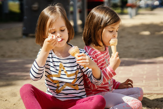 two little girls eats ice-cream while sitting on the playground in the summer