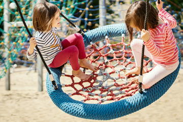 two little girls are swinging on a swing at the playground on a summer day - obrazy, fototapety, plakaty