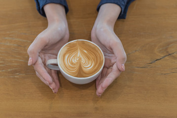 Female hands holding a cup of coffee with foam over wooden table, top view