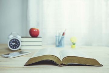 Holy bible book with smartphone, alarm clock, stack of books, apple and some accessory on wooden table with window light, Christian background, copy space