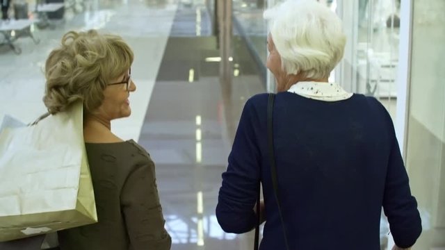 Tilt up medium shot of two elderly women talking to each other and carrying paper bags with purchases when walking through shopping center, rear view