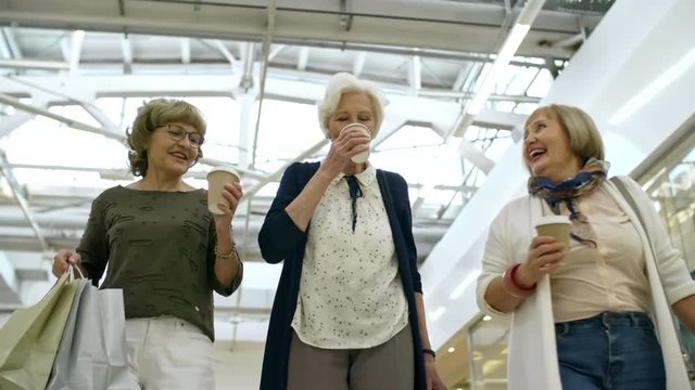 Group of three happy retired women walking along shopping mall, drinking takeaway coffee and talking to each other after buying new clothes