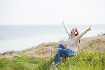 Young screaming fun pretty woman in casual clothes with headphones listening music sitting on grass spreading hands pointing index fingers up near water on green field background. Lifestyle concept.