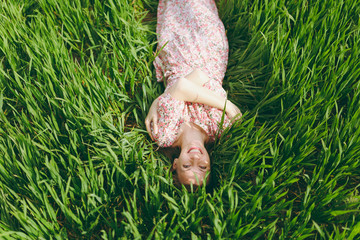 Young smiling woman in light patterned dress lying on grass holding hands folded looking up resting in sunny weather in field on bright green background. Spring nature. Lifestyle, leisure concept.
