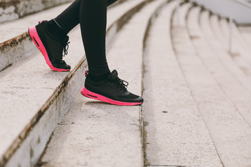 Fototapeta premium Cropped photo close up of female legs in sportswear, black and pink woman sneakers doing sport exercises, going down on stairs outdoors. Fitness, healthy lifestyle concept. Copy space advertisement.