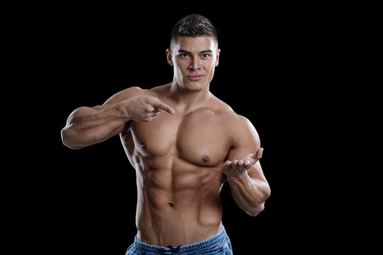 A smiling male bodybuilder points to his open palm. The picture is suitable for advertising sports goods and services. Isolated in black background.