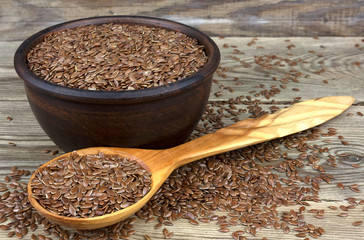 Flax seeds in bowl with scattered grains on wooden background with wooden spoon.