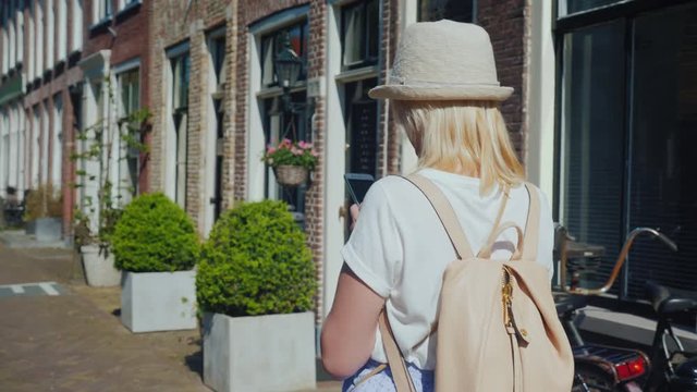 A woman in a hat and with a backpack walks along the narrow street of the ancient city of Delft in the Netherlands. Uses a smartphone. Tourism in Europe concept