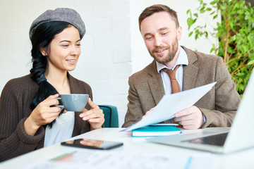 Portrait of two contemporary business people, man and woman, holding documents while discussing strategy during meeting in cafe, both smiling cheerfully