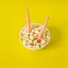 die Beine des Puppenmädchens in der Platte mit Popcorn auf gelbem Hintergrund. Kreatives Sommerkonzept des Kinos © EkaterinaChebotareva