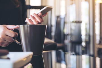 Closeup image of a woman holding and using smartphone while drinking coffee in cafe