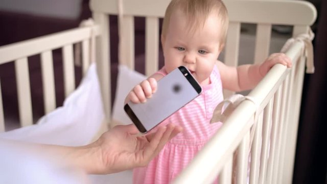 Cute baby in crib touch smartphone. Child with mobile phone in crib. Female hand give infant touch screen mobile. Baby technology concept