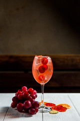 A photo of an orange cold summer alcoholic cocktail in a wine glass, full of ice cubes, garnished with red grapes and dried citrus. White wooden table.