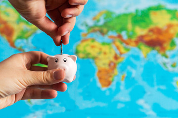 A tiny piggy bank is held in the hand. A colorful map of the world in the background. Travel...