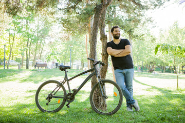 Fototapeta na wymiar Smiling man leaning on a tree in the park with his bicycle next to him