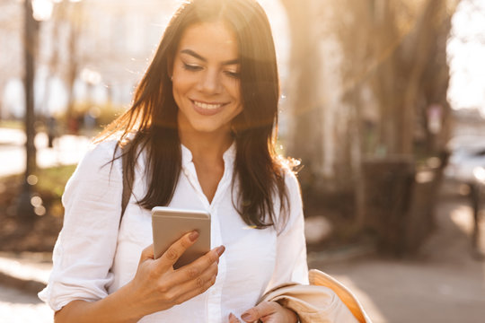 Image of Smiling brunette woman in shirt using her smartphone
