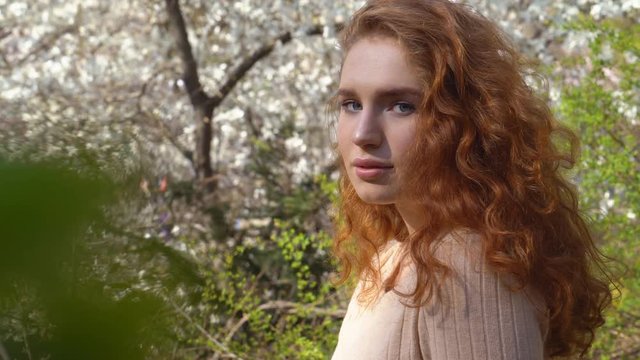 female portrait close-up. funny girl with natural red hair and blue eyes. woman tourist laughs in the botanical garden. in the background a blooming pink and white magnolia garden. spring freshness of