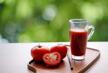 Tomato juice in glass and fresh tomatoes on wooden tray with blurred bokeh background from green tree in garden, healthy concept.