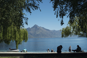Personas descansando a la orilla de un lago de agua clara y azul frente a un paisaje de montañas...