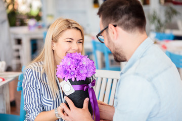 Young man giving beautiful flower bouquet to his girlfriend for birthday or anniversary. Romantic moments in a cafe