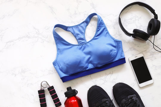Top view of flat lay fitness equipment with sport bra, sneakers shoes, smartphone, earphones and bottle of water on white marble background