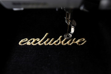 embroidery of gold lettering "exclusive" on black velvetely fabric with embroidery machine - front view with part of machine - background and foreground blanked out blurry