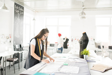 Wide angle portrait of focused Asian woman tracing patterns for custom made clothes while standing at tailors table and working in modern design atelier with people working in background, copy space