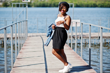 Fototapeta na wymiar Stylish african american model in glasses hat, jeans jacket and black skirt posed outdoor on pier against lake.