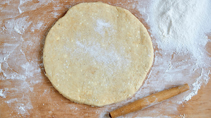 Raw round dough for pizza or pie with rolling pin and scattered flour on the wooden kitchen table. Baking background. Top view