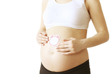 Close up of pregnant woman wearing white supportive seamless maternity bra & black yoga pants with blank alarm clock shaped sticker on big bare belly. Expectancy concept. Background, copy space.