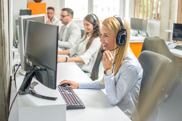 Call center operators.Young beautiful business woman with headset working on computer in office with colleagues in the background.