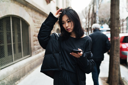 Beautiful asian woman in stylish outfit readjusting the sunglasses on her head while walking the street with a mobile phone in her hand. Hipster girl chatting online while going down the city.