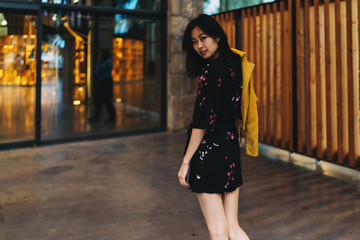 Young asian woman wearing mini dress looking back at the camera on her way to the shopping center. Beautiful tourist female from Japan with long dark hair standing beside the enter to the museum.