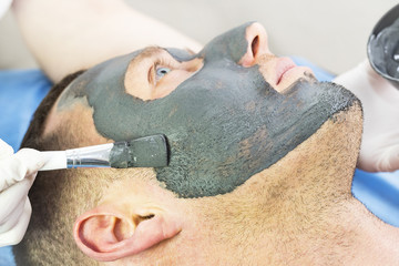 A man does the procedure cleaning his face with a clay mask in the beauty salon 