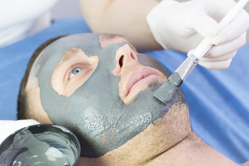 A man does the procedure cleaning his face with a clay mask in the beauty salon 