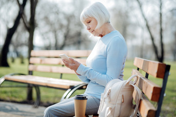 Great gadget. Concentrated aged woman typing on her phone while sitting on the bench