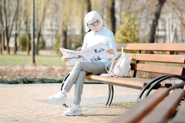 Staying informed. Serious aged woman reading a newspaper while sitting on the bench