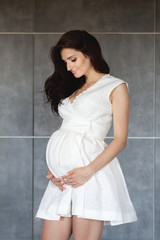 A pregnant brunette in a white dress embraced her pregnant belly and looked at him. Gray background