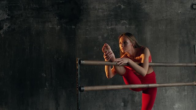 Female athlete is pulling her leg and back during training in gym. She is leaning on a barre by leg and tipping forward