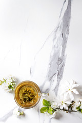 Glass cup of hot green tea with spring flowers white magnolia and cherry blooming branches over white marble texture background. Top view, copy space.